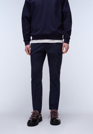 Mann trägt marineblaue Cargohosen, schwarze klobige Schuhe mit pinken Schnürsenkeln, marineblauen Pullover und hellgraues Hemd vor einem schlichten Hintergrund.