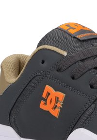 Czarno-brązowe buty sportowe z teksturowanym wierzchem, okrągłymi sznurówkami i pomarańczowym logo DC z boku. Widoczna biała gumowa podeszwa.