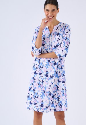 Vrouw die lacht met een hand bij haar mond, gekleed in een bloemenjurk tot op de knie in pastelblauwe, roze en witte tinten, staand tegen een effen achtergrond.