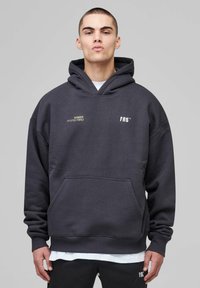 Man wearing dark gray hoodie with front pocket and white text on chest, standing against plain light gray background.