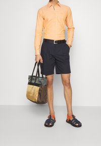 Hombre que lleva una camisa de manga larga naranja, pantalones cortos azul marino, cinturón negro y sandalias azules y naranjas, sosteniendo una bolsa de mano multicolor en una mano.