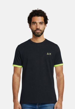 Uomo con capelli ricci e barba indossa una maglietta nera con bordi delle maniche verde lime e piccoli simboli gialli a forma di triangolo e X sul petto.