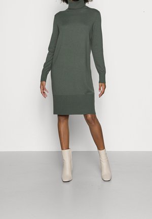 Frau trägt ein langärmeliges, grünes Rollkragen-Strickkleid kombiniert mit off-white Knöchelstiefeln und steht vor einem einfarbigen hellen Hintergrund.