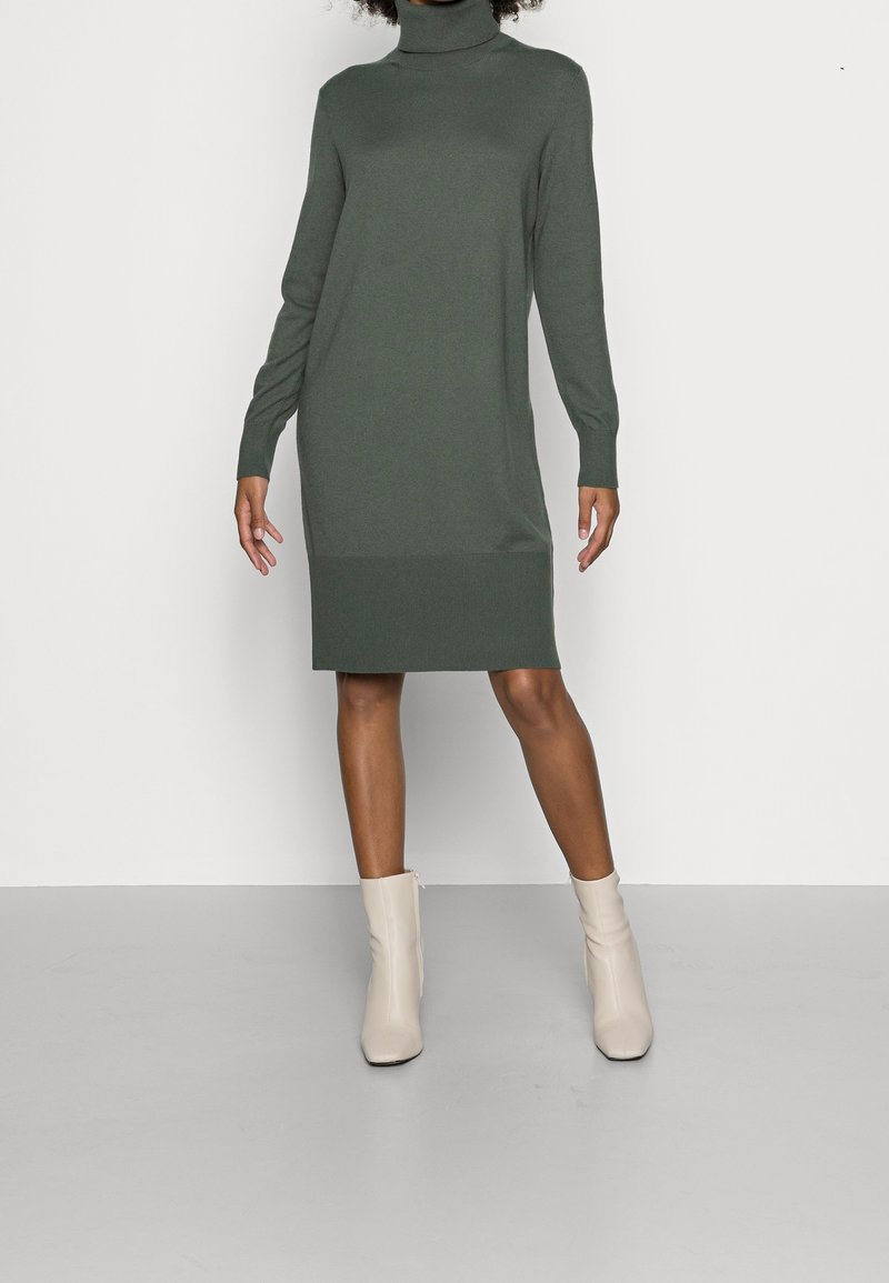 Femme portant une robe-pull verte à col roulé manches longues, associée à des bottines écru, debout devant un fond clair uni.