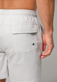 Costumi da bagno a strisce bianche e grigie con vita elasticizzata, tasca posteriore con pattina e un'etichetta con il marchio, che mostrano un tessuto testurizzato.