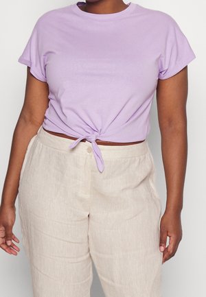 Personne portant un haut à manches courtes violet clair noué à la taille et un pantalon beige taille haute sur un fond uni.