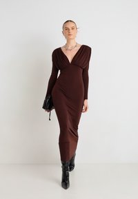 Καφέ φόρεμα με μακριά μανίκια και βαθύ V-λαιμόκοψη, εφαρμοστό και με ραντιές στους ώμους, συνδυασμένο με μαύρες μπότες μέχρι το γόνατο.