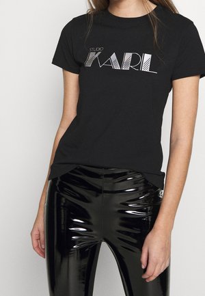 Camiseta negra de manga corta con el texto gráfico en blanco "STUDIO KARL". Combinada con pantalones negros de tiro alto y brillo, destacando el contraste de texturas.