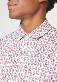 Chemise blanche à boutons avec un motif répété de flamants roses. Le col est traditionnel, et le tissu semble léger et lisse.