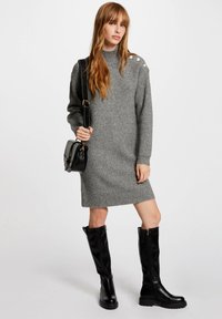 Abito in maglia grigia con collo alto e texture a coste. Abbinato a stivali neri al ginocchio con cerniere dorate e una borsa strutturata.