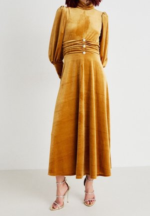Frau trägt ein senfgelbes Samt-Midikleid mit Puffärmeln, einem gerafften Taillenbereich mit drei Knöpfen und goldenen Riemchen-High Heels.