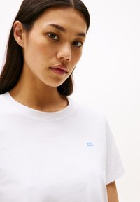 Weißes Baumwoll-T-Shirt mit einem runden Halsausschnitt und kurzen Ärmeln, ausgestattet mit einem kleinen blauen Logo auf der Brust. Glatte Textur.