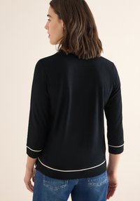 Un haut en jersey noir avec des manches trois-quarts, un passepoil blanc le long des manches et de l'ourlet, et une coupe décontractée. Porté avec un jean bleu.