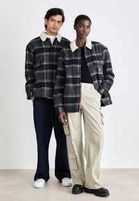 Flanelljacken mit Schwarz-Grau-Karomuster, ausgestattet mit weißen Sherpakragern. Ein Modell trägt beige Cargo-Hosen, das andere marineblaue Hosen.