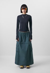 Haut noir à manches longues en tissu texturé ; jupe maxi verte avec taille élastique et poches latérales ; portée avec des chaussures jaunes et des lunettes de soleil réfléchissantes.