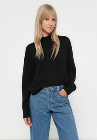 Svart strikkegenser med høy hals, avslappet passform og lange ermer, kombinert med lyseblå vide jeans. En enkel, teksturert design.