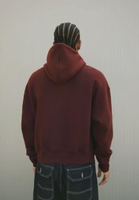 Persona con trecce a cornrow che indossa un felpa color bordeaux e pantaloni blu scuro con tasche posteriori, in piedi contro un muro grigio chiaro.