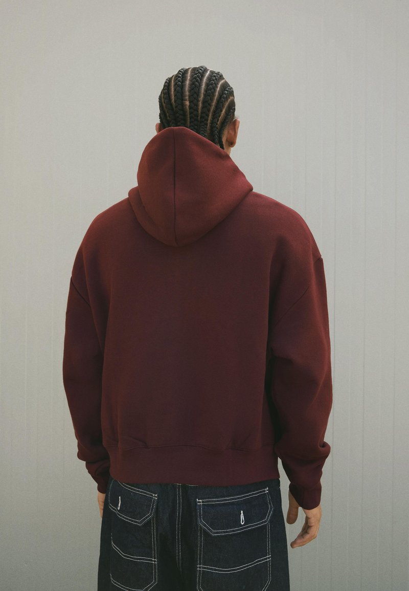 Oseba s cornrow pletenicami, oblečena v maroon hušo in temno modre hlače z zadnjimi žepi, stoji ob svetlo sivi steni.