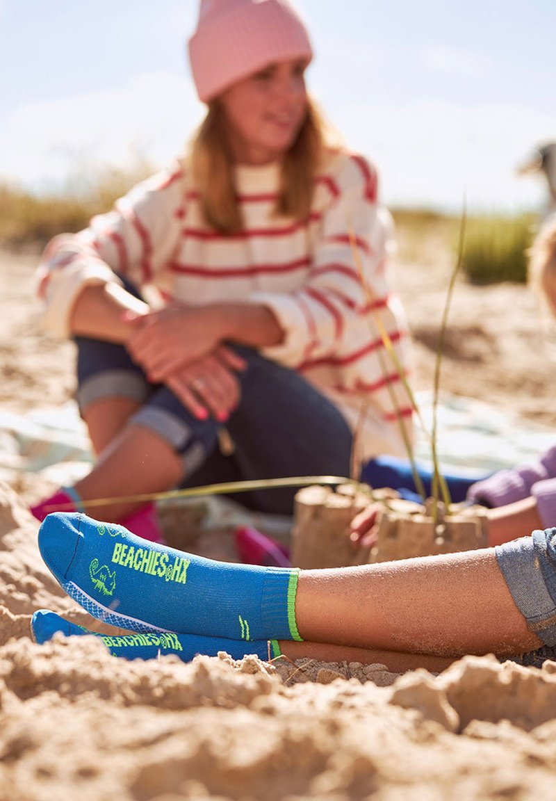 Person sitzt im Sand und trägt blaue Socken mit der Aufschrift "BEACHES, MA". Im Hintergrund ist eine andere Person verschwommen zu sehen, die eine pinke Mütze und einen gestreiften Pullover trägt.