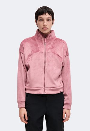 Persoon draagt een roze jas met hoge kraag en rits, met gestructureerde stof op schouders en mouwen en een zwarte broek, kijkt recht vooruit met een neutrale uitdrukking.