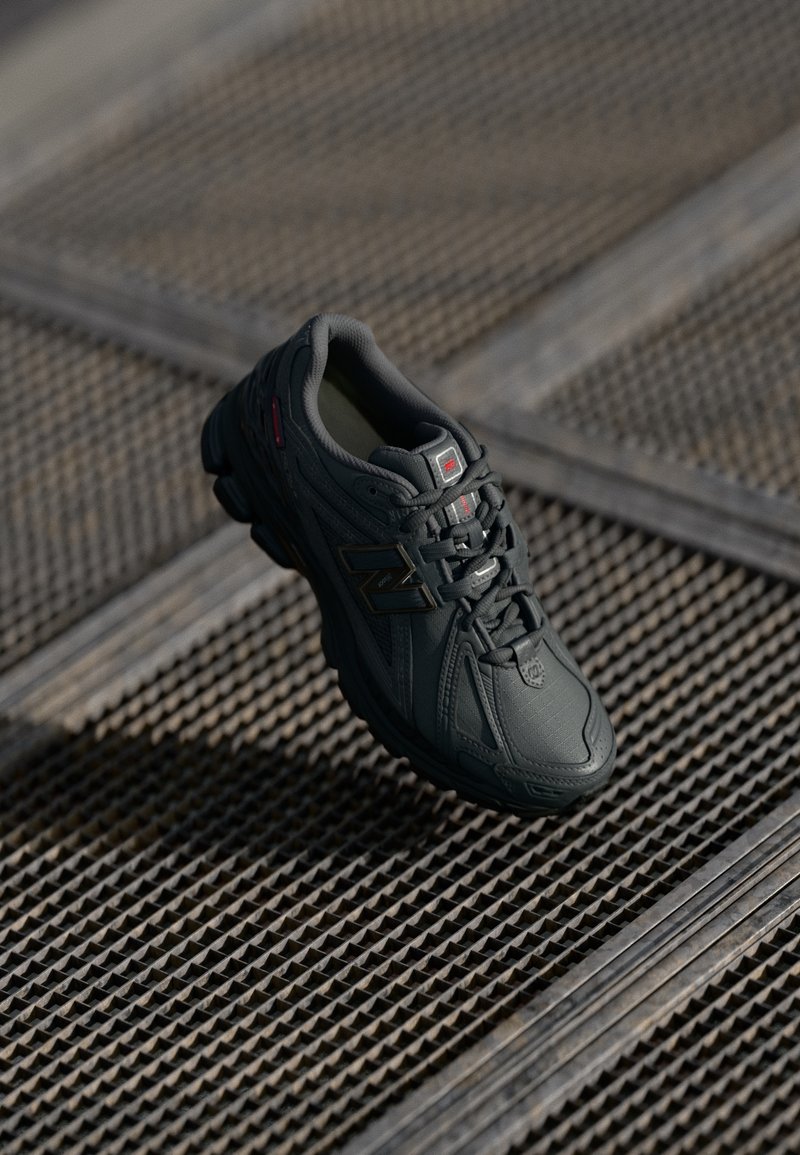 Baskets New Balance noires avec un design texturé et des lacets, reposant sur un sol en grille métallique avec des lignes diagonales et des ombres.