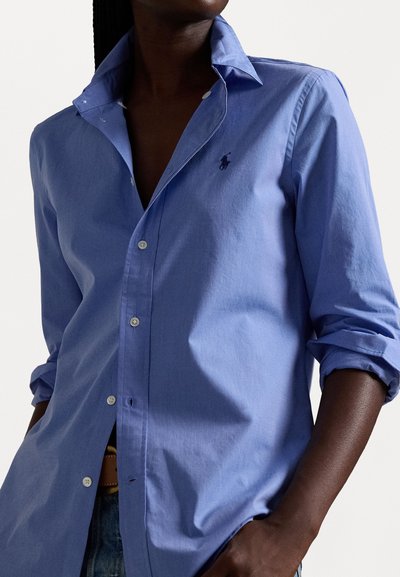 Chemise bleu clair à manches courtes avec un col classique, présentant un petit logo brodé et des manches retroussées. Texture lisse, coupe décontractée.