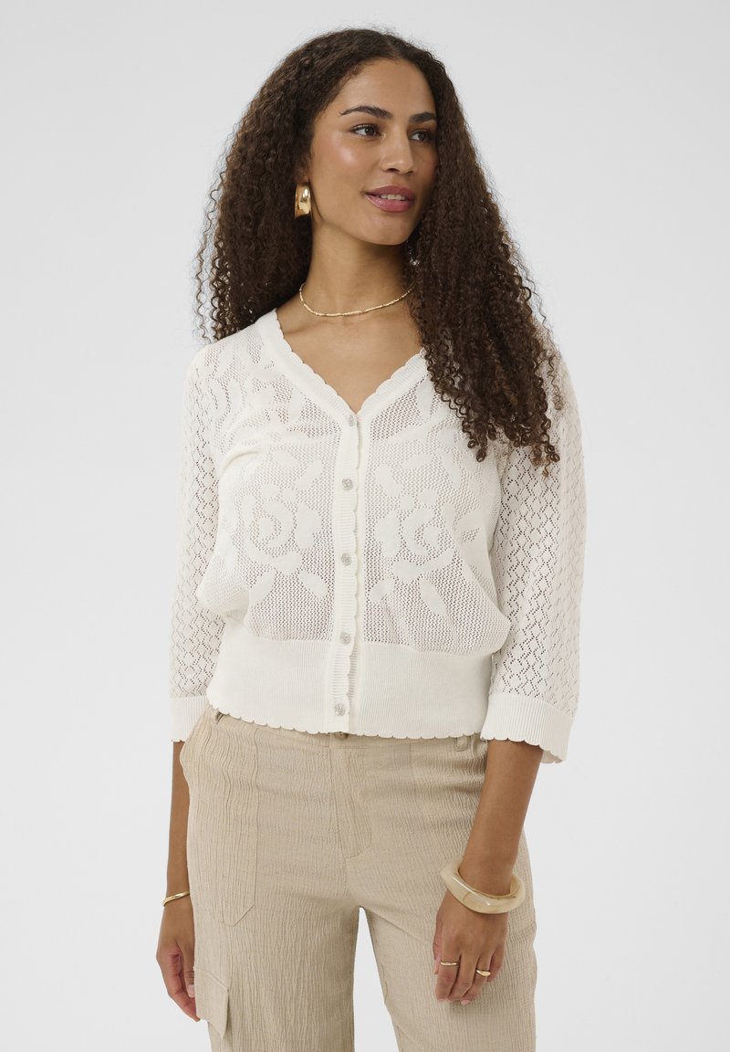 Femme aux longs cheveux bouclés portant un cardigan en maille blanc à motifs floraux et un pantalon beige, accessoirisée avec des boucles d'oreilles, un collier et des bracelets en or.