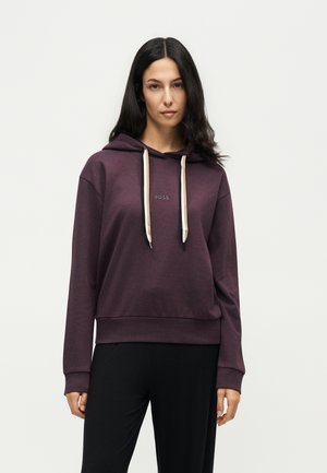 Vrouw met lang donker haar, gekleed in een donkerpaarse hoodie met crèmekleurige trekkoorden en een zwarte broek, staand tegen een effen lichte achtergrond.