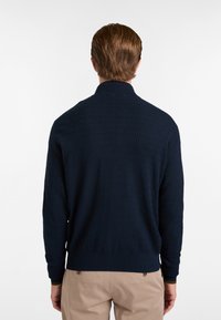 Jersey azul marino con un patrón de zigzag texturizado, cuello y puños y dobladillo de canalé, mostrado por la parte de atrás contra un fondo claro.