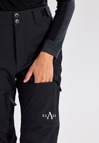 Zwarte softshellbroek met een ritszak aan de zijkant, klittenbandsluiting en een wit logo. Materiaal lijkt glad en heeft een slim fit design.