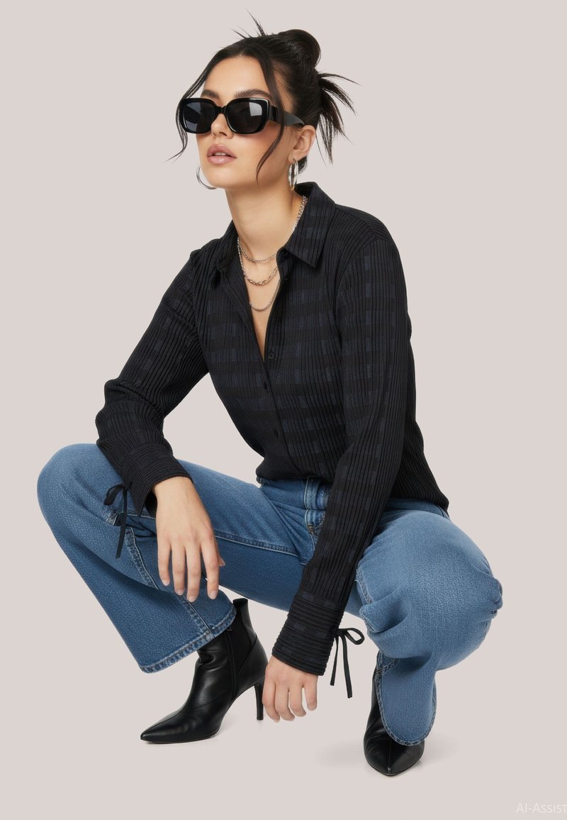 Femme portant des lunettes de soleil noires, une chemise noire boutonnée, un jean bleu et des bottes noires à talons, accroupie sur un fond clair.