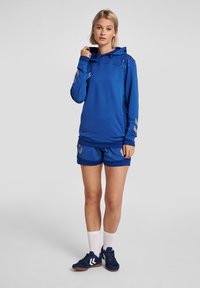 Felpa sportiva blu con parte superiore a fantasia, abbinata a pantaloni corti blu. Presenta dettagli in argento e tasche laterali. Include scarpe scure e calze bianche.