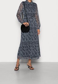 Longue robe bleue et noire à imprimé zèbre, manches longues en voile transparent, taille cintrée et silhouette fluide, accompagnée d'un petit sac noir.