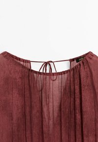 Blouse bordeaux transparente avec encolure froncée et fermeture par lien fin au dos, montrant la texture légère du tissu et l'étiquette de la marque à l'intérieur.