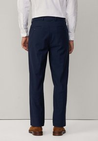 Pantalon bleu marine à coupe droite, avec deux poches arrière à boutons, porté avec une chemise blanche à manches longues et des chaussures marron.