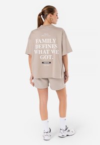 Beige oversized t-skjorte med hvit tekst på baksiden, kombinert med matchende shorts, hvite sokker og sneakers. Glatt bomullsstoff.