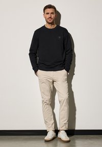 Svart crew-neck sweatshirt, beige chinos och vita sneakers med mockadetaljer. Modellen står med händerna i fickorna mot en neutral bakgrund.