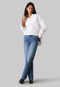 Top sin mangas blanco acanalado, camisa blanca de gran tamaño y jeans rectos azules. Botas negras de tacón. Tejido suave, diseño casual.
