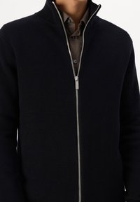 Lätt svart zip-up-tröja med ribbad textur, hög krage och silverdragkedjedetalj. Bärs över en brun skjorta med knäppning.