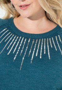 Türkiser Strickpullover mit rundem Halsausschnitt und silbernen Pailletten, die im Sonnenstrahlenmuster an der oberen Vorderseite angeordnet sind.