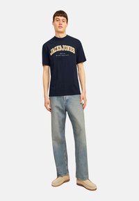 Navy blue cotton t-shirt with "JACK&JONES" and "EST. 1990" graphics. Paired with light blue denim jeans and beige shoes.