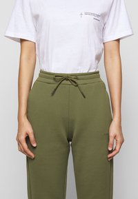 Olivgrüne Sweatpants mit einem Kordelzug-Bund und Seiten pockets, kombiniert mit einem weißen Baumwoll-T-Shirt, das bedruckten Text auf der Brust trägt.