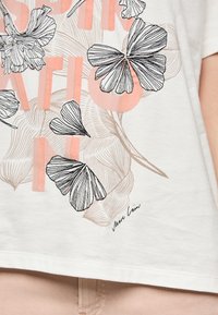 White cotton t-shirt featuring black floral line drawings and soft pink accents, with the text "INSPIRATION" partially visible and a designer signature.