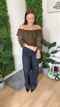 Bluza olive cu umerii goi și detaliu de volane, combinată cu blugi largi de culoare închisă și tocuri negre ascuțite, pe fundal de frunziș verde.