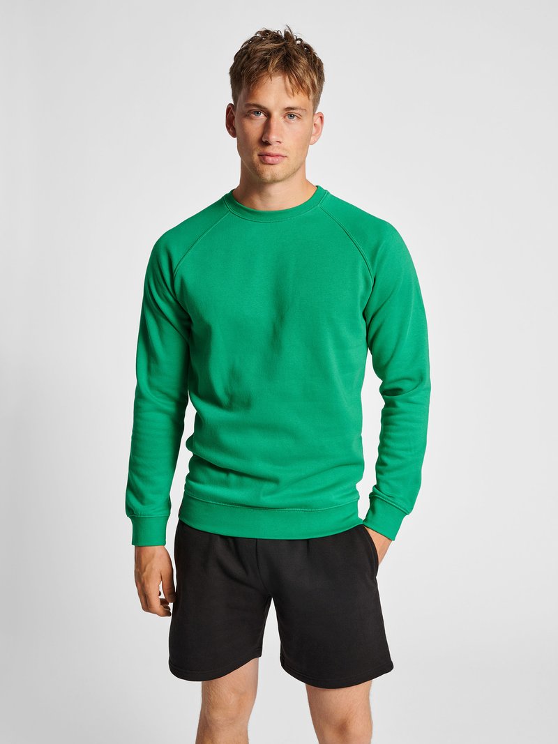 Junger Mann steht, trägt ein leuchtend grünes Sweatshirt mit langen Ärmeln und schwarze Shorts vor einem schlichten hellgrauen Hintergrund.