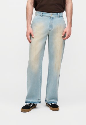 Άτομο που φοράει ανοιχτό μπλε τζιν με φαρδιά πόδια και μπεζ λεκέδες, και μαύρα αθλητικά παπούτσια με καφέ σόλες, στέκεται μπροστά σε απλό φόντο.