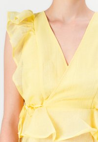 Top de lino amarillo con mangas con volantes, escote en pico profundo y un detalle de lazo en la cintura, con una textura ligera y ligeramente transparente.