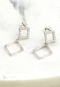 Silverhängande örhängen har en fyrkantig sten med klara rhinestones och en hängande öppen fyrkant nedanför. Slät textur, geometrisk design.