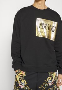Svart sweatshirt med en glänsande guldlogo som visar "VERSACE JEANS COUTURE" och "PIECE NO." Tyget ser mjukt ut med en avslappnad passform.