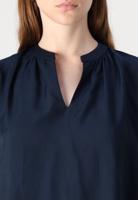Blusa blu navy con scollatura a V e dettagli arricciati sul collo. Realizzata in tessuto liscio, con maniche corte.